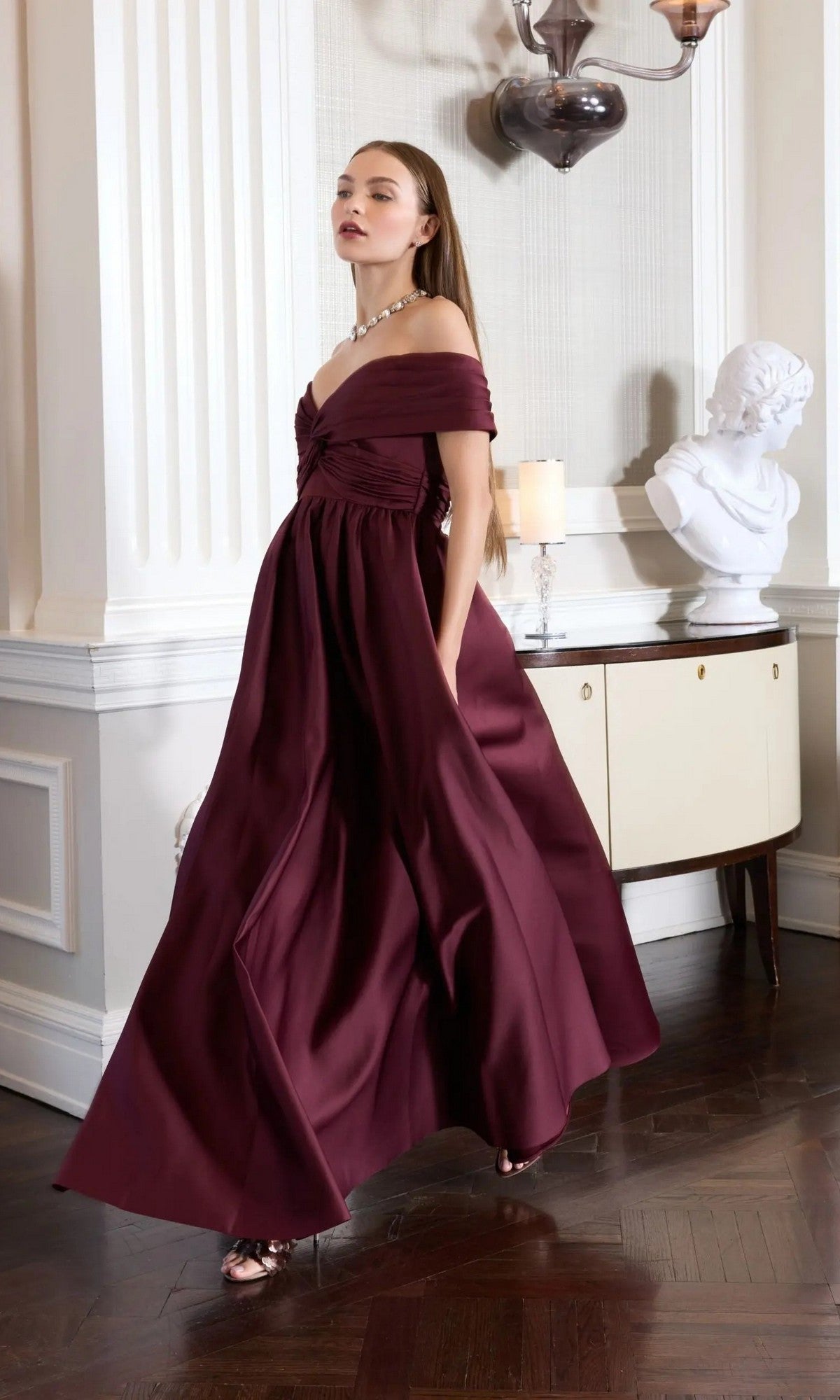 A woman in the Mac Duggal 56188 Long Formal Dress stands indoors near a white bust sculpture and a cabinet with a lit candle, surrounded by elegant decor.